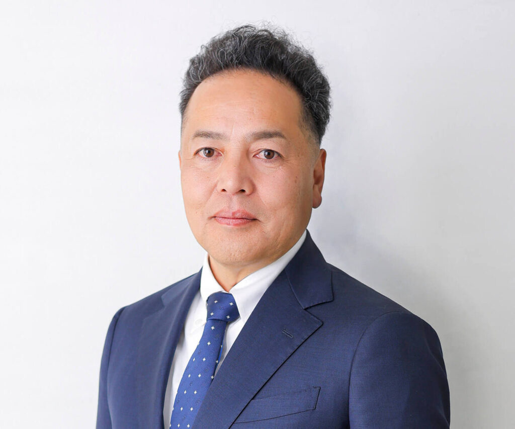 白い背景の前で、青いスーツと白いシャツ、青いネクタイを着用した男性が、正面を向いて落ち着いた表情で写っているビジネス用ポートレート写真。短く整えた黒髪と淡い笑みが特徴。